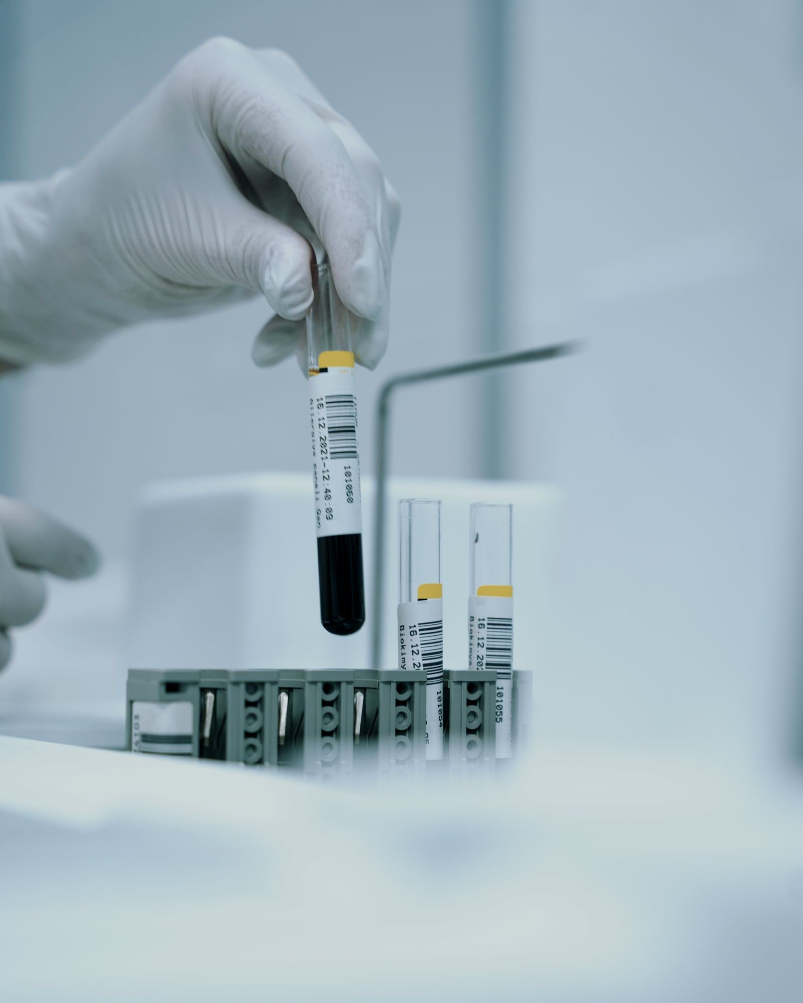 Gloved hand lifting a barcode-labelled sample tube from a rack in a laboratory.